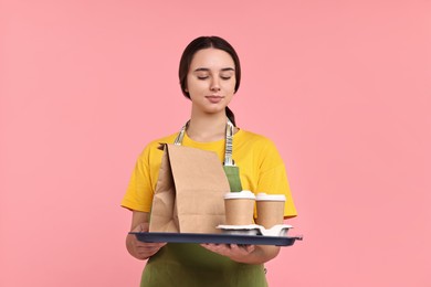 Girl in apron holding takeaway cups of coffee and paper bag on pink background. Work for teenagers