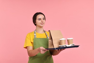 Photo of Girl in apron holding takeaway cups of coffee and paper bag on pink background. Work for teenagers