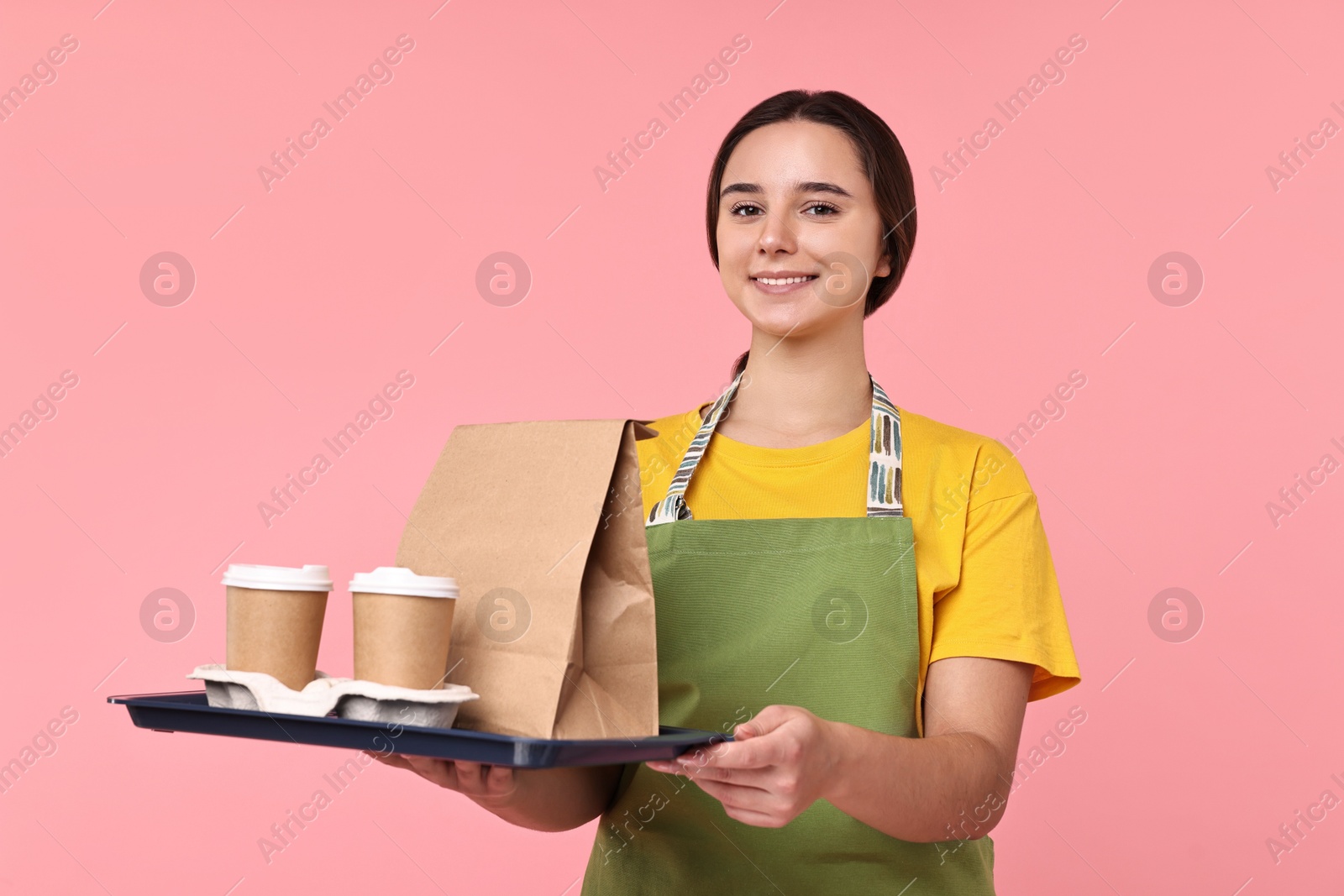 Photo of Girl in apron with takeaway cups of coffee and paper bag on pink background. Work for teenagers