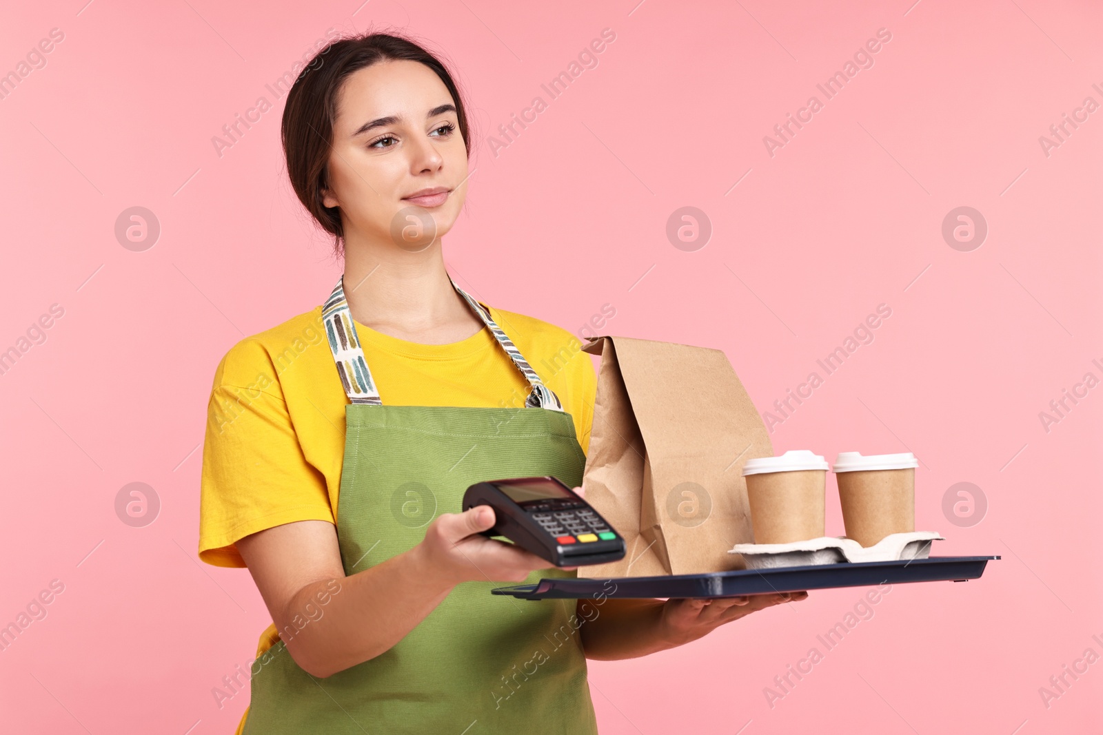 Photo of Girl in apron with takeaway cups of coffee, paper bag and payment terminal on pink background. Work for teenagers