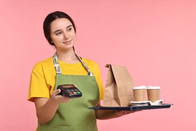 Photo of Girl in apron with takeaway cups of coffee, paper bag and payment terminal on pink background. Work for teenagers