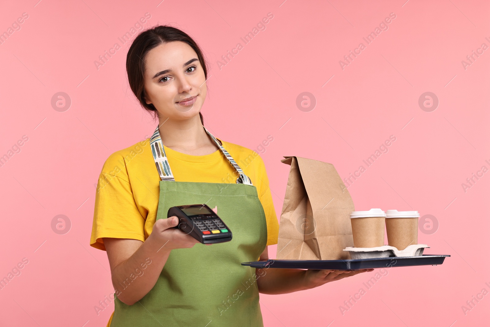 Photo of Girl in apron with takeaway cups of coffee, paper bag and payment terminal on pink background. Work for teenagers