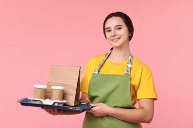 Photo of Girl in apron with takeaway cups of coffee and paper bag on pink background. Work for teenagers