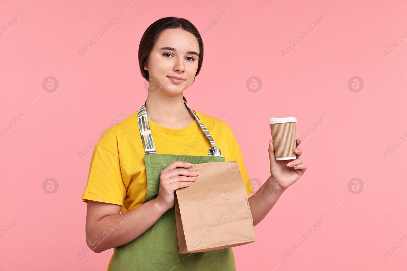 Photo of Girl in apron holding takeaway cup of coffee and paper bag on pink background. Work for teenagers