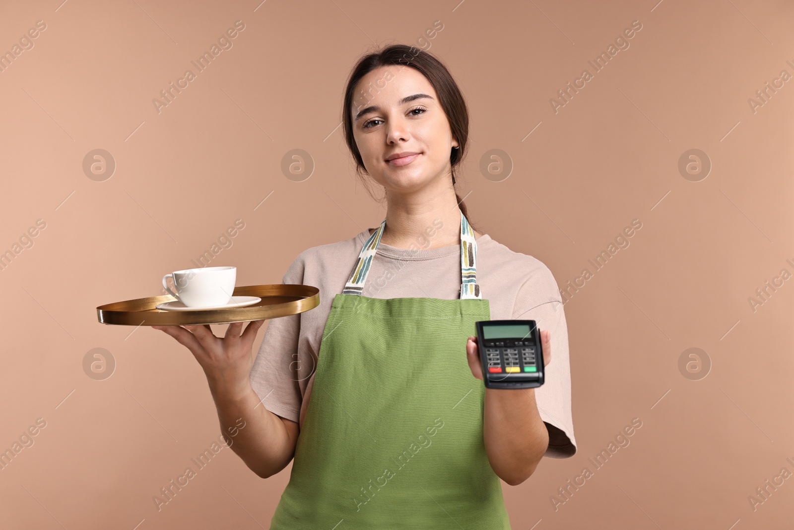 Photo of Girl in apron with cup of coffee and payment terminal on pale brown background. Work for teenagers