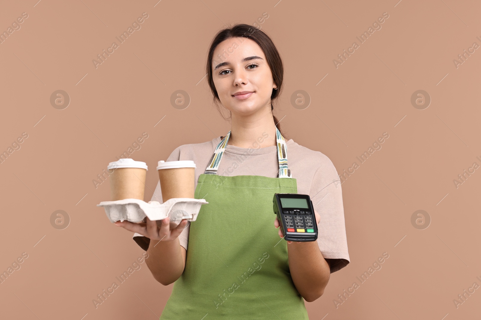 Photo of Girl in apron with takeaway paper cups of coffee and payment terminal on pale brown background. Work for teenagers