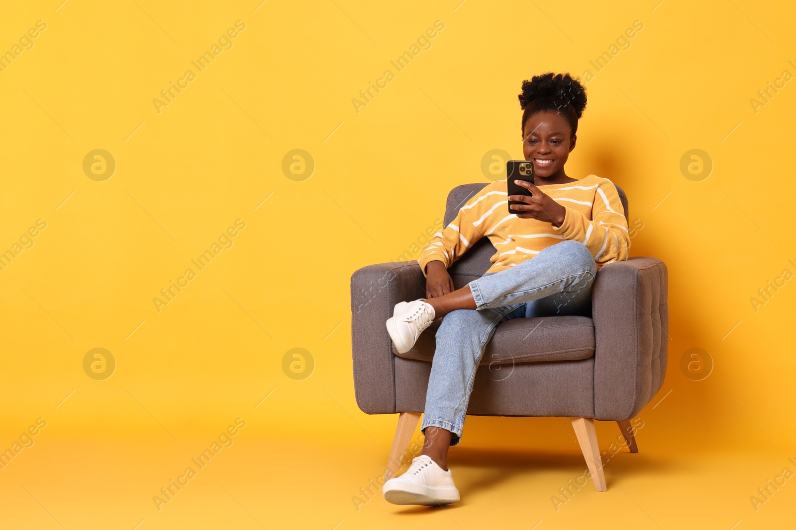 Photo of Smiling woman with smartphone sitting in armchair on yellow background. Space for text