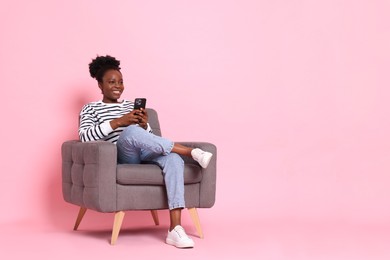 Smiling woman with smartphone sitting in armchair on pink background. Space for text