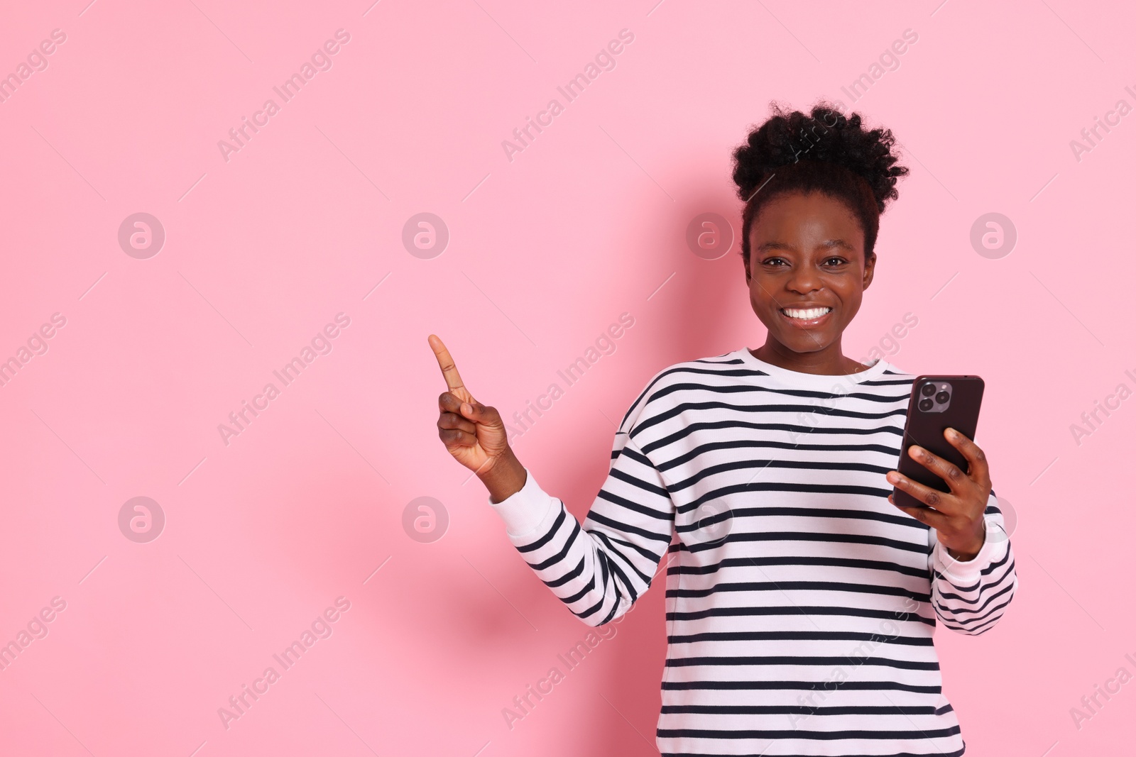 Photo of Cheerful woman with smartphone pointing at something on pink background. Space for text