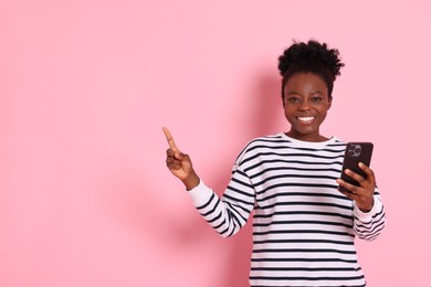 Photo of Cheerful woman with smartphone pointing at something on pink background. Space for text