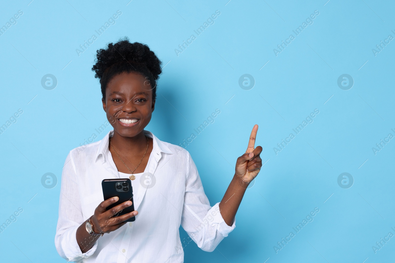 Photo of Cheerful woman with smartphone pointing at something on light blue background. Space for text