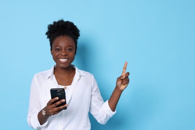 Photo of Cheerful woman with smartphone pointing at something on light blue background. Space for text