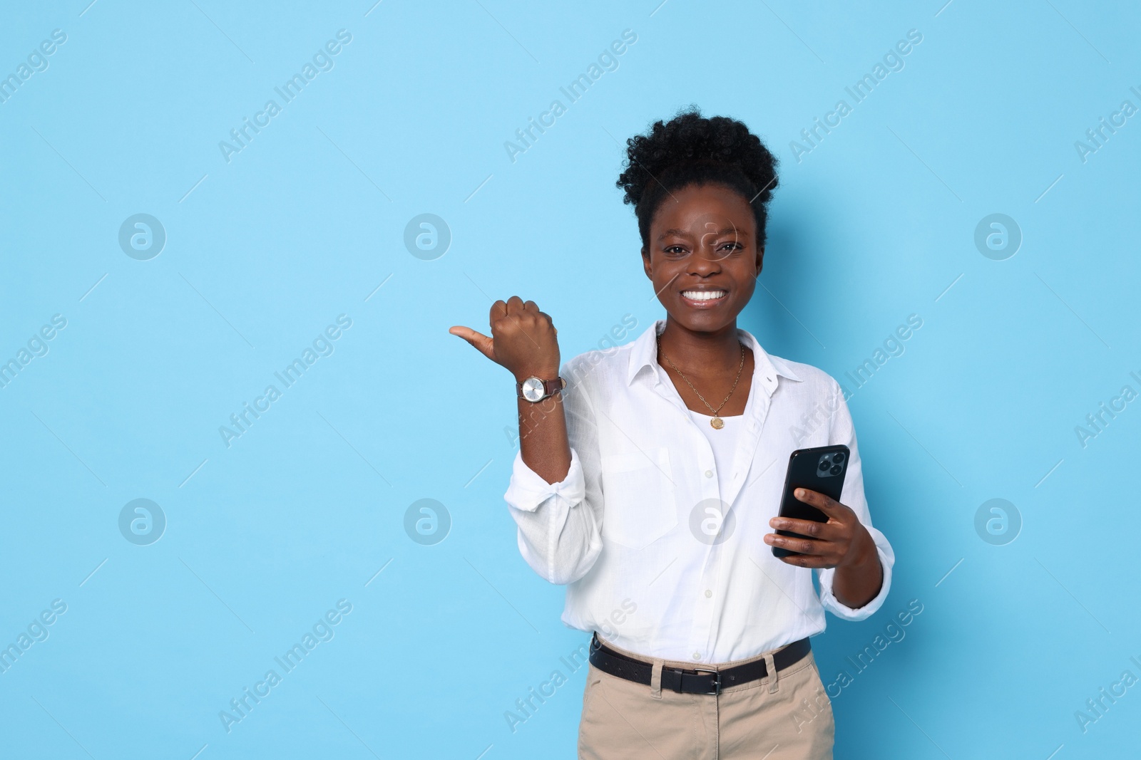 Photo of Cheerful woman with smartphone pointing at something on light blue background. Space for text