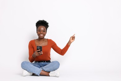 Cheerful woman with smartphone pointing at something on white background. Space for text