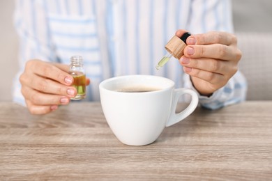 Photo of Young woman putting CBD tincture into cup with drink at wooden table, closeup