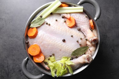 Photo of Whole raw turkey, cut carrot, celery and spices in pot on grey textured table, top view