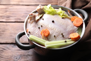 Photo of Whole raw turkey, cut carrot, celery and spices in pot on wooden table, closeup