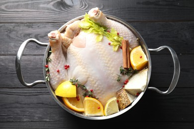 Photo of Whole raw turkey, citrus fruits and spices in pot on black wooden table, top view