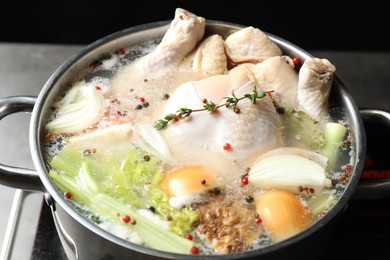 Photo of Whole fresh turkey, cut onion, celery and spices boiling in pot on table against black background, closeup