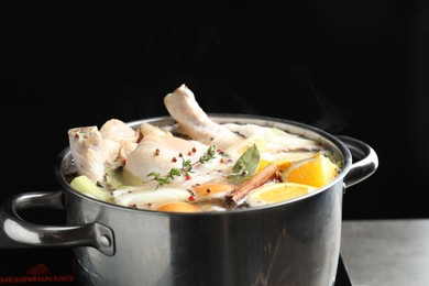 Photo of Whole fresh turkey, cut citrus fruits, thyme and spices boiling in pot on table against black background, closeup