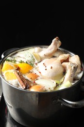 Photo of Whole fresh turkey, cut citrus fruits, thyme and spices boiling in pot on black background, closeup