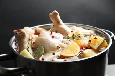 Photo of Whole fresh turkey, cut citrus fruits and spices boiling in pot on black background, closeup