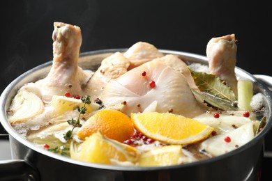 Photo of Whole fresh turkey, cut citrus fruits and spices boiling in pot on black background, closeup