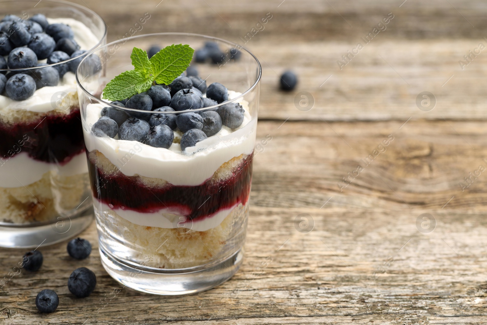 Photo of Tasty trifle dessert. Sponge cake, blueberries, jam and whipped cream in glasses on wooden table, closeup. Space for text