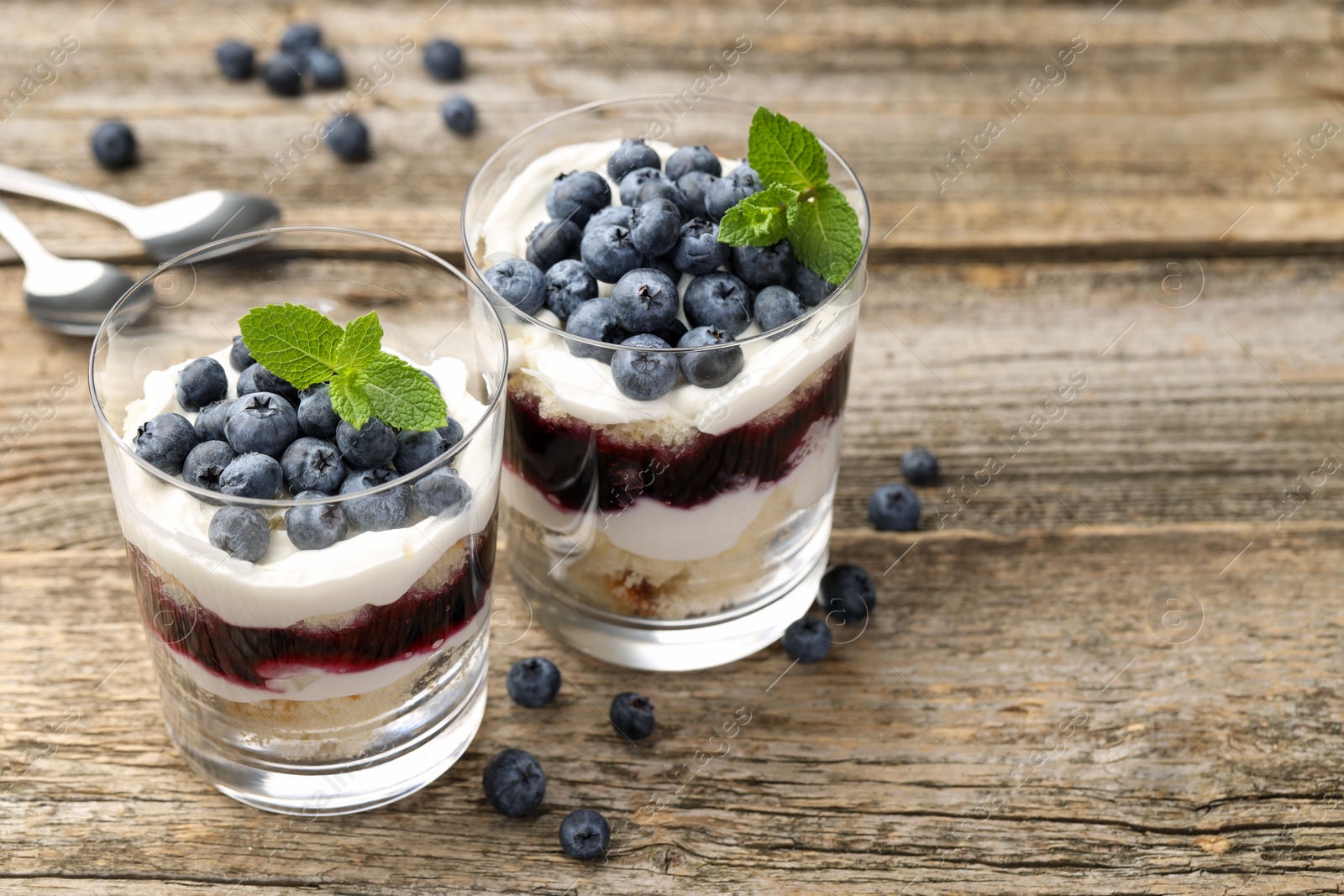 Photo of Tasty trifle dessert. Sponge cake, blueberries, jam and whipped cream in glasses on wooden table, closeup. Space for text