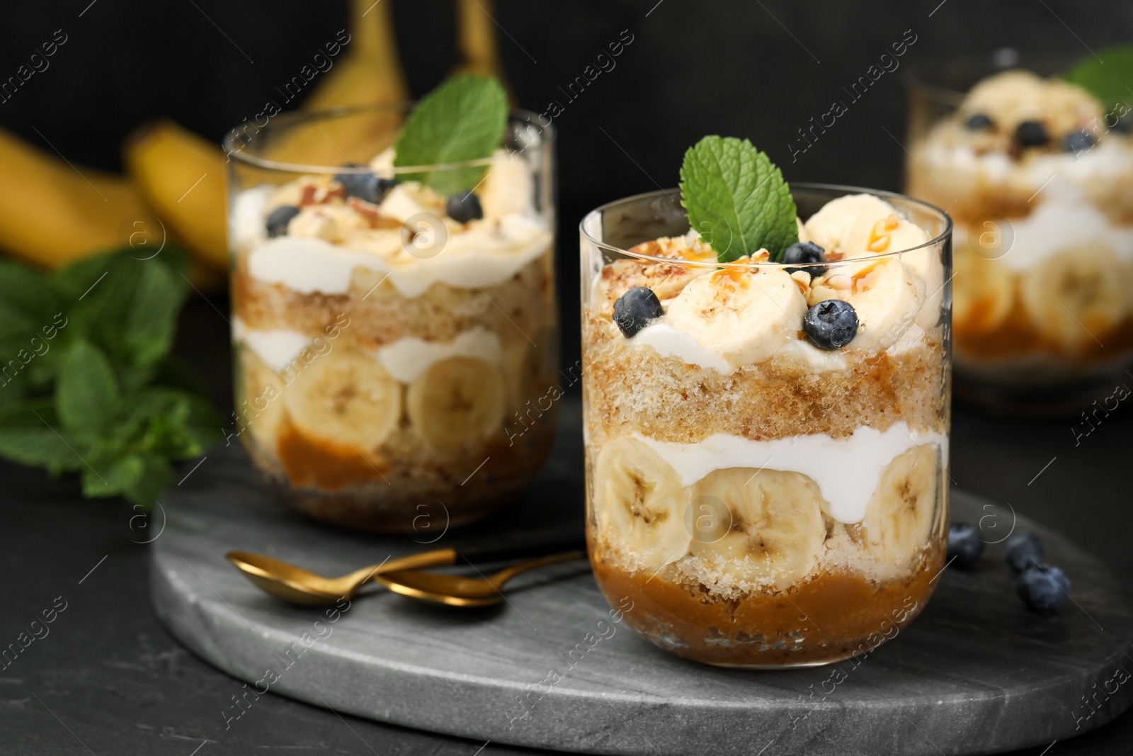 Photo of Tasty trifle dessert. Sponge cake, blueberries, banana and whipped cream in glasses on black table, closeup