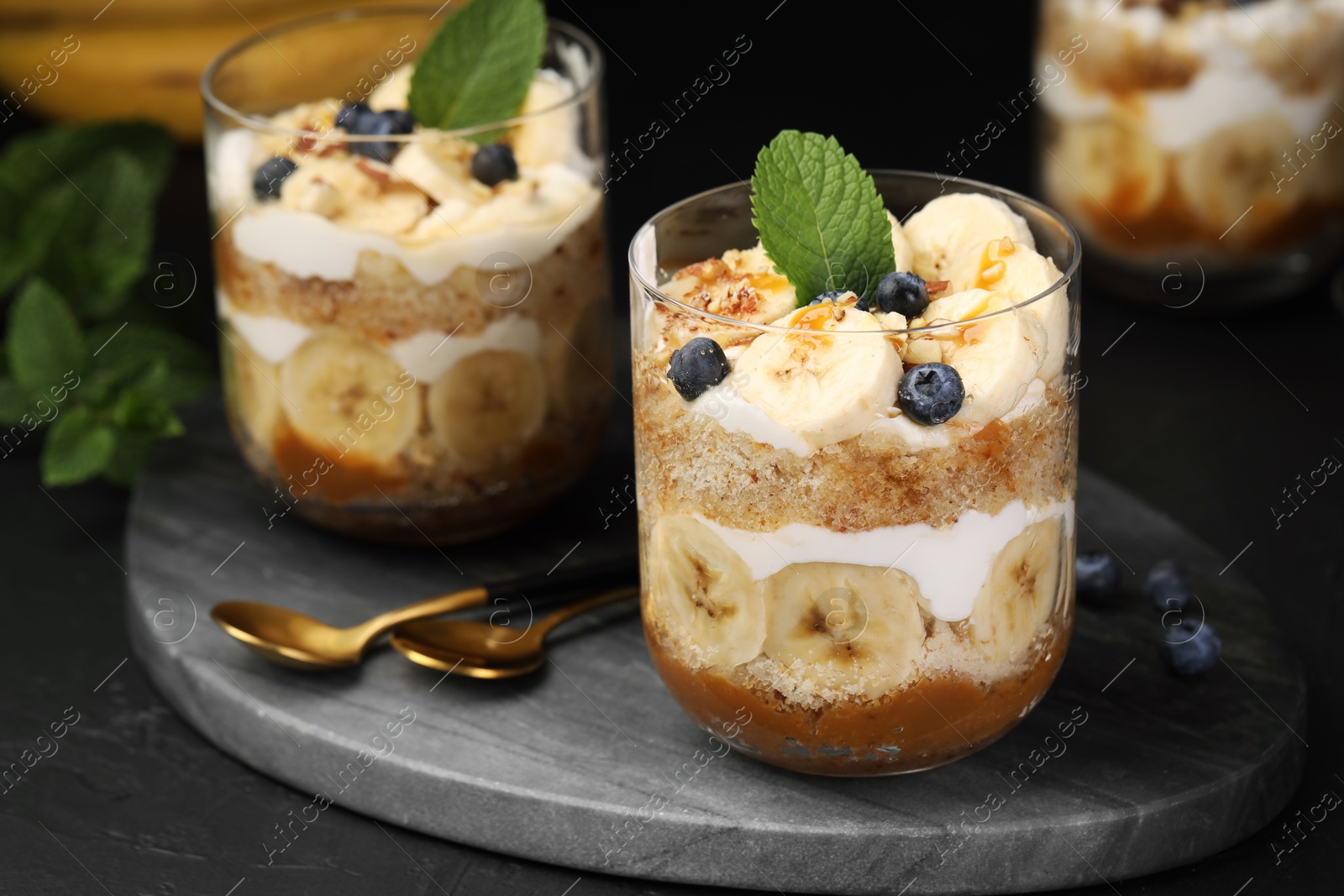 Photo of Tasty trifle dessert. Sponge cake, blueberries, banana and whipped cream in glasses on black table, closeup