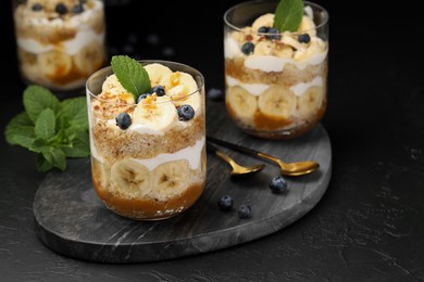 Photo of Tasty trifle dessert. Sponge cake, blueberries, banana and whipped cream in glasses on black table