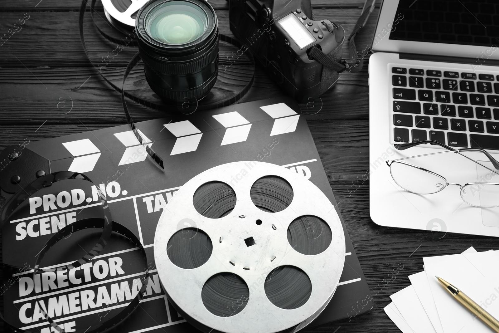 Photo of Movie clapper, film reel, laptop and camera lens on black wooden table, above view