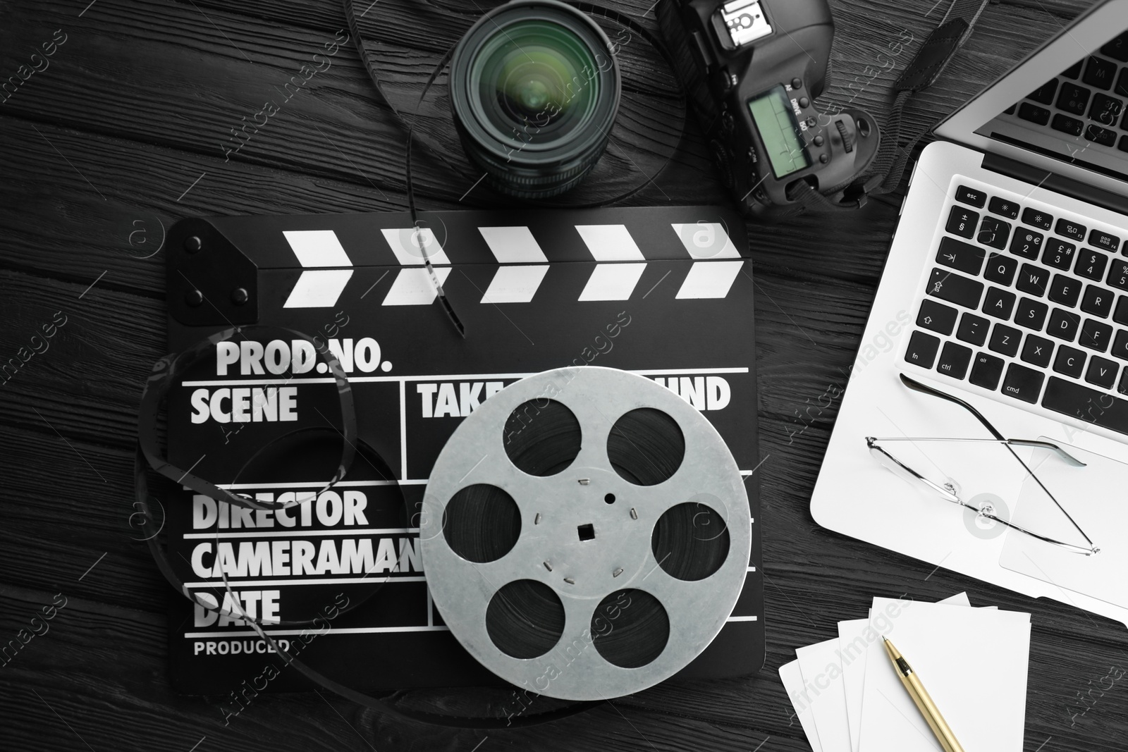 Photo of Movie clapper, film reel, laptop and camera lens on black wooden table, flat lay