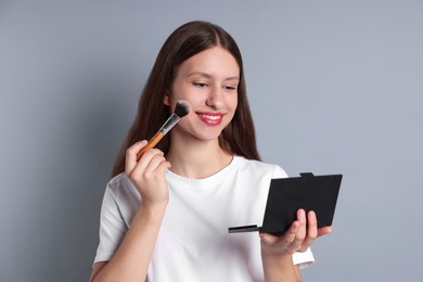 Photo of Smiling teenage girl applying blusher with makeup brush on grey background