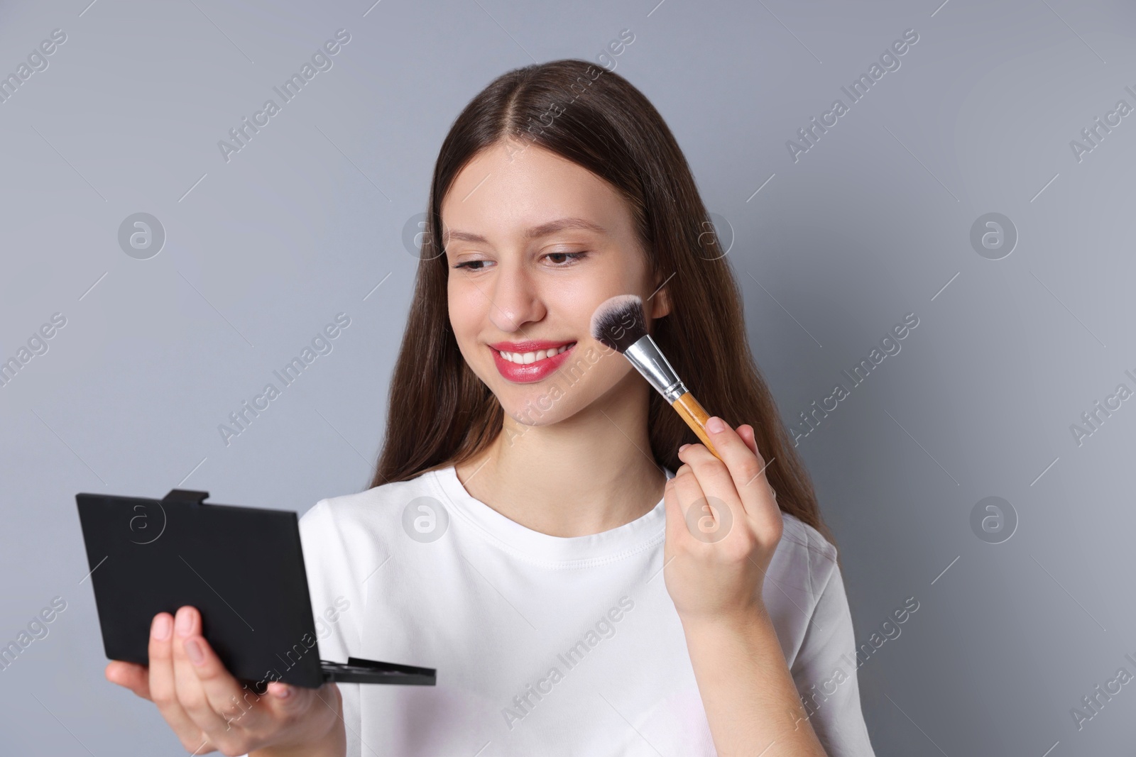 Photo of Smiling teenage girl applying blusher with makeup brush on grey background