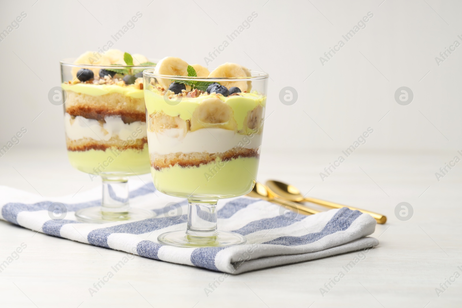 Photo of Tasty trifle dessert. Banana, blueberries, sponge cake and whipped cream in glasses on white wooden table, space for text