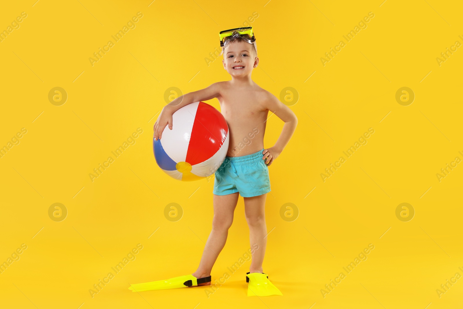 Photo of Cute little boy in beachwear with diving mask and inflatable ball on orange background