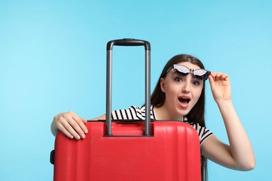 Emotional woman with suitcase wearing sunglasses on light blue background