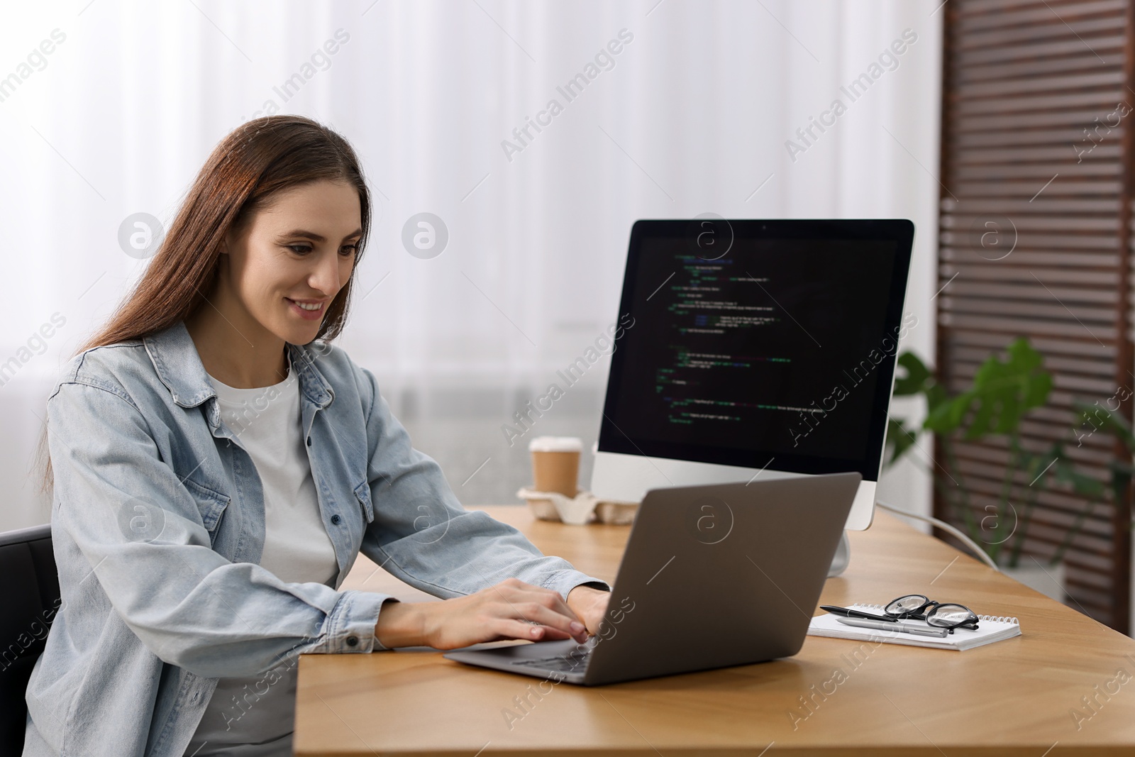 Photo of Programmer working on laptop and computer at wooden desk indoors