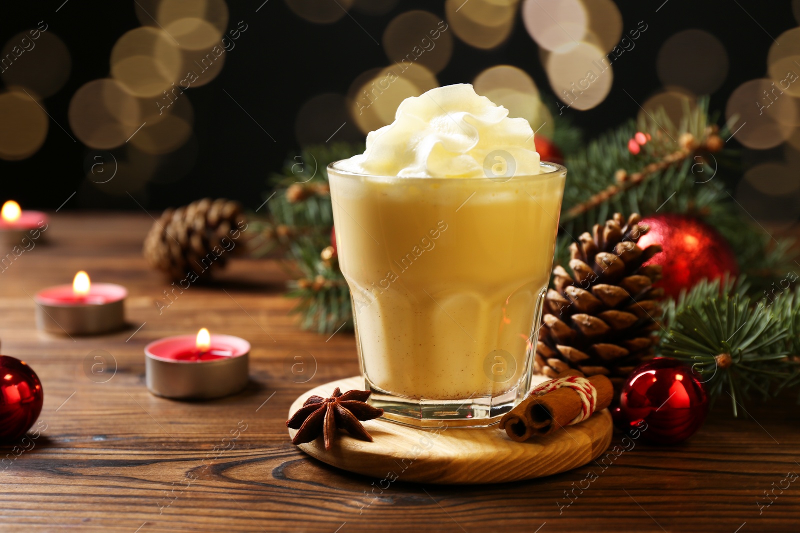 Photo of Tasty Christmas cocktail (eggnog) in glass, festive decor and burning candles on wooden table, closeup