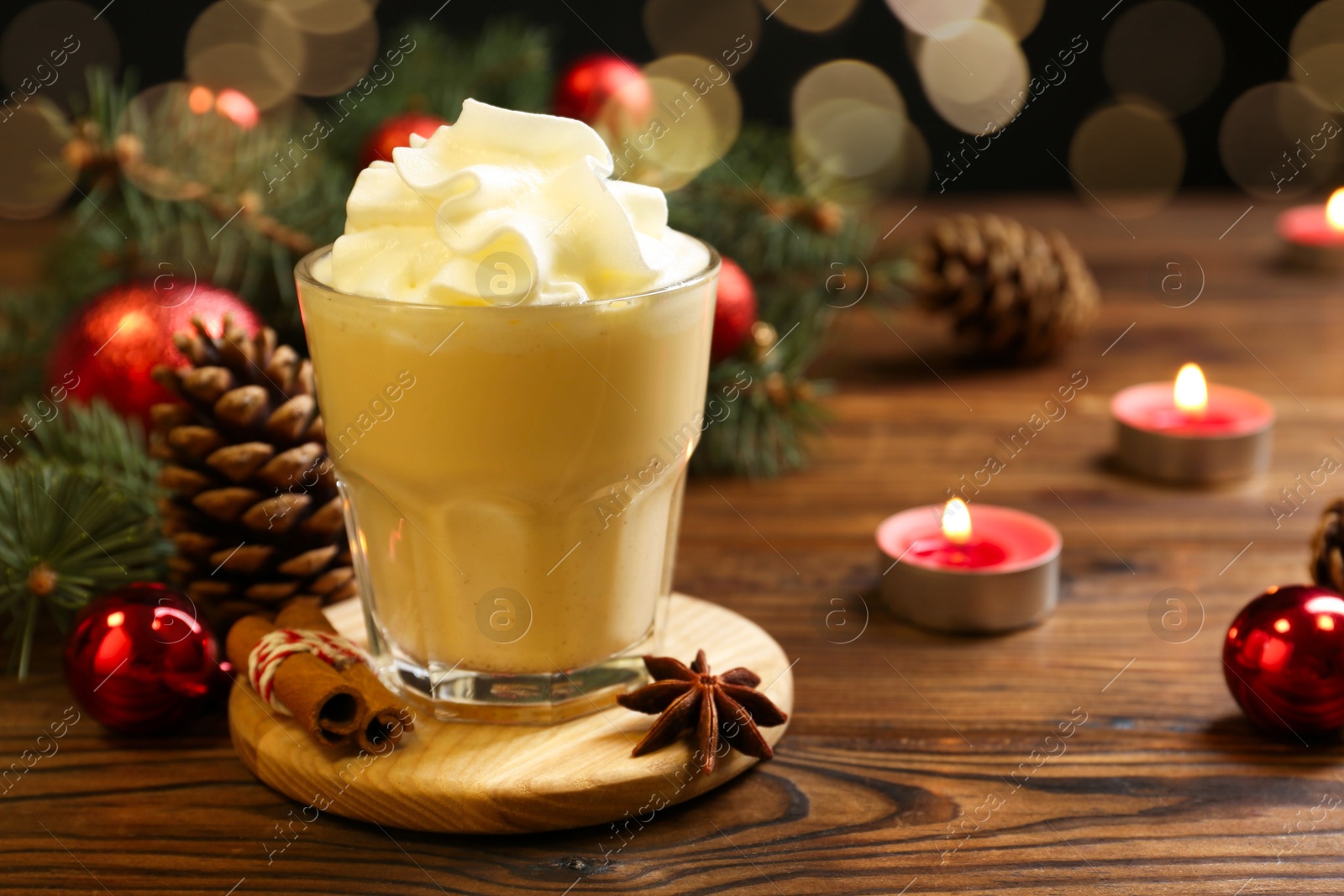 Photo of Tasty Christmas cocktail (eggnog) in glass, festive decor and burning candles on wooden table, closeup