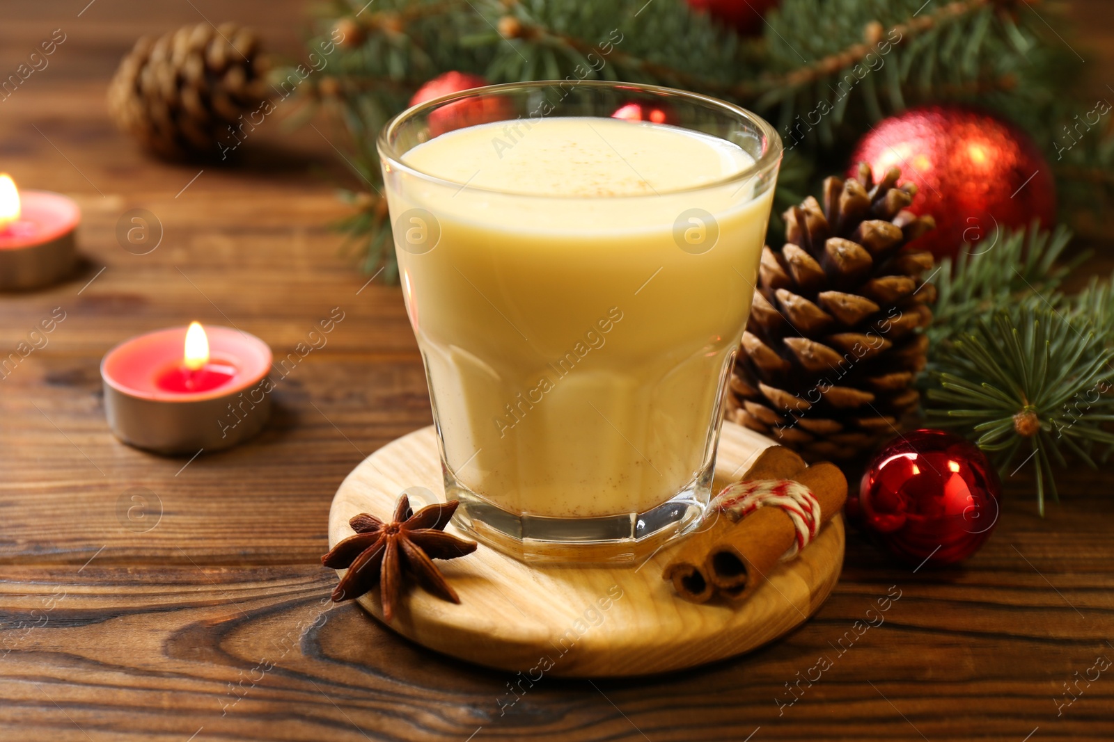 Photo of Tasty Christmas cocktail (eggnog) in glass, festive decor and burning candles on wooden table, closeup