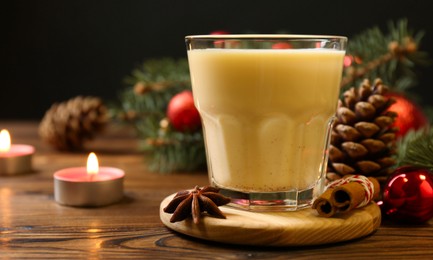 Photo of Tasty Christmas cocktail (eggnog) in glass, festive decor and burning candles on wooden table, closeup