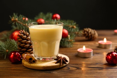 Photo of Tasty Christmas cocktail (eggnog) in glass, festive decor and burning candles on wooden table, closeup