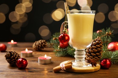 Photo of Tasty Christmas cocktail (eggnog) in glass, festive decor and burning candles on wooden table