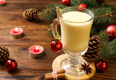 Photo of Tasty Christmas cocktail (eggnog) in glass, festive decor and burning candles on wooden table, closeup