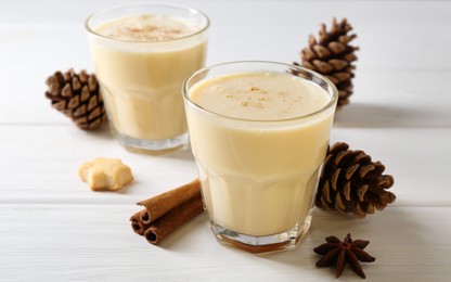 Photo of Tasty Christmas cocktail (eggnog) in glasses, pine cones and spices on white wooden table, closeup