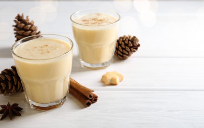 Photo of Tasty Christmas cocktail (eggnog) in glasses, pine cones and spices on white wooden table, closeup. Space for text