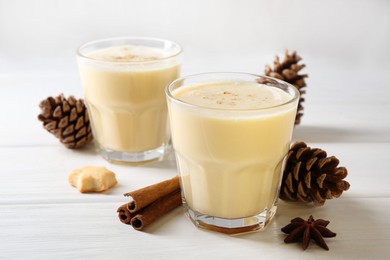 Photo of Tasty Christmas cocktail (eggnog) in glasses, pine cones and spices on white wooden table, closeup